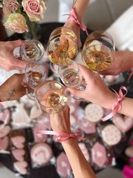 Top-down view of a champagne toast at a bridal shower — hands tied with pink ribbons hold wine and champagne glasses over a table of pink heart-shaped decorated cookies and pale roses