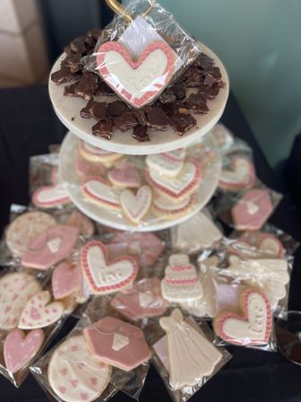 Tiered dessert display of individually wrapped pink-and-white iced heart and dress-shaped sugar cookies with chocolate bark on a marble stand, cute party favors for a bridal shower or romantic celebration