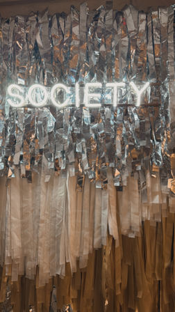 Neon "SOCIETY" sign atop shimmering silver fringe and cream-gold streamers — festive photo booth backdrop.