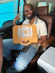 Smiling passenger in a shuttle van flashing a peace sign and holding an orange luggage-shaped Cancun airport transfer sign, relaxed travel vibe