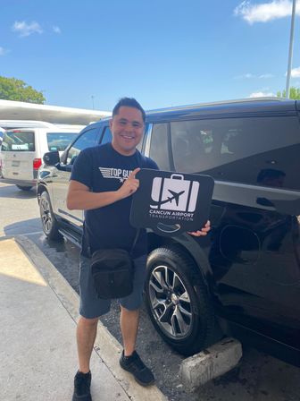 Smiling traveler holding an airport-transfer sign with a luggage and airplane icon beside a black SUV in a sunny airport pickup area.