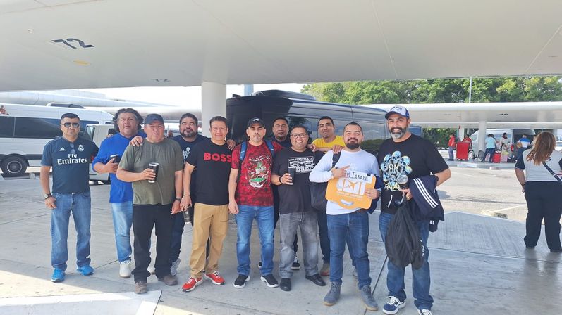 Smiling group of casually dressed men at an airport curbside pickup area under a canopy, standing in front of shuttle vans with luggage and coffee — travel group at an airport terminal.