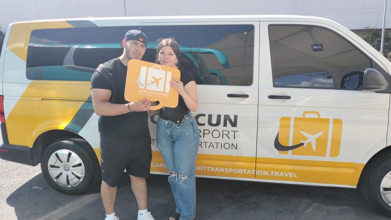 Two travelers holding an orange airport sign standing beside a white-and-yellow Cancun airport transfer van