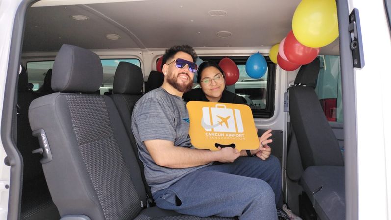 Two smiling passengers in a decorated shuttle van holding a bright orange Cancun airport shuttle sign with colorful balloons