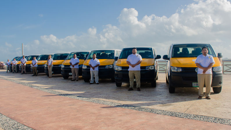 Line of yellow-and-white shuttle vans parked along a Cancún waterfront promenade with uniformed drivers standing in front under a bright blue sky