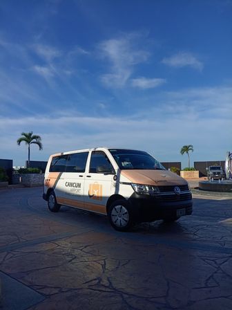 White-and-orange airport shuttle van parked on a stamped stone driveway with palm trees and clear blue sky in Cancun, Mexico