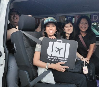 Four smiling travelers inside a white shuttle van holding a luggage sign with an airplane icon, posing for a vacation airport transfer photo.