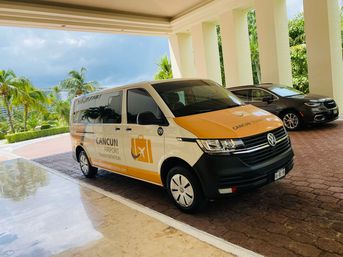 Yellow-and-white airport shuttle van parked under a hotel porte-cochère at a Cancún resort, palm trees and cloudy sky in the background.