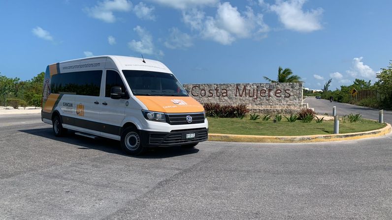 White-and-orange shuttle van parked by a stone 'Costa Mujeres' entrance sign on a sunny day in Costa Mujeres, Cancun, Mexico