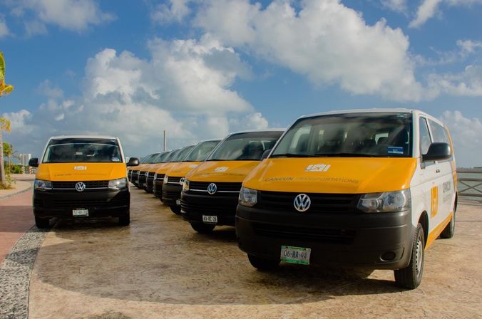 Row of yellow-and-white shuttle vans lined up on a sunny Cancún waterfront promenade with palm trees and blue sky dotted with clouds