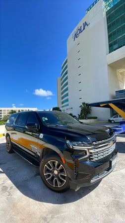 Shiny black full-size SUV parked at a sunny hotel drop-off beside palm trees and a modern white building with blue-tinted windows under a clear blue sky.