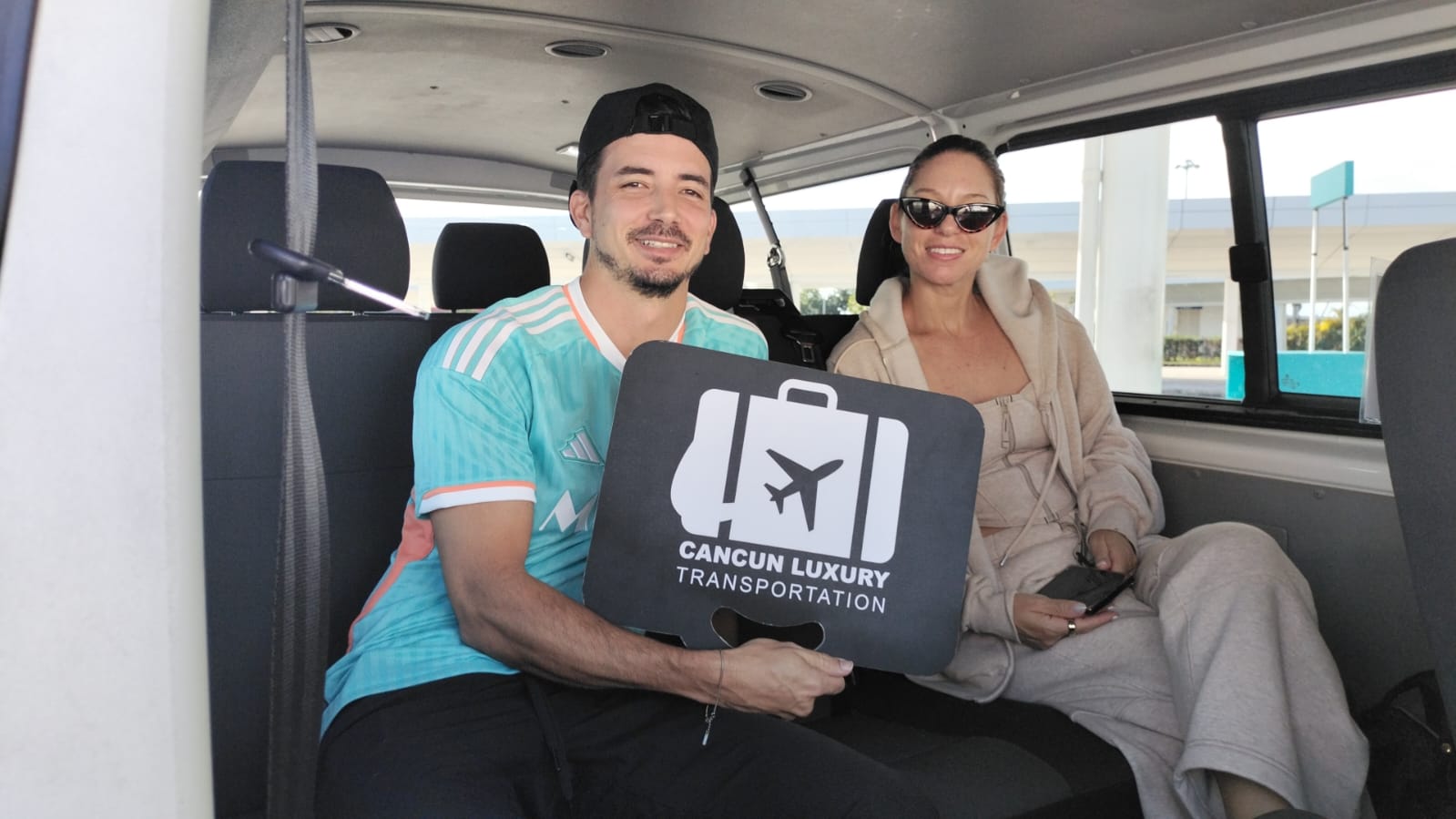 Two smiling travelers seated in a Cancun shuttle van holding a black suitcase-shaped airport transfer sign with an airplane icon