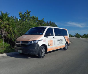 Peach-and-white airport shuttle van parked on a sunny coastal road near palm trees, branded for Cancun airport transportation under a clear blue sky.
