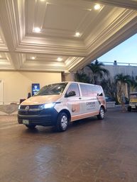 Orange-and-white Volkswagen airport shuttle van marked "Cancun" parked under a hotel porte-cochere at dusk, with palm trees and illuminated coffered ceiling in the background.