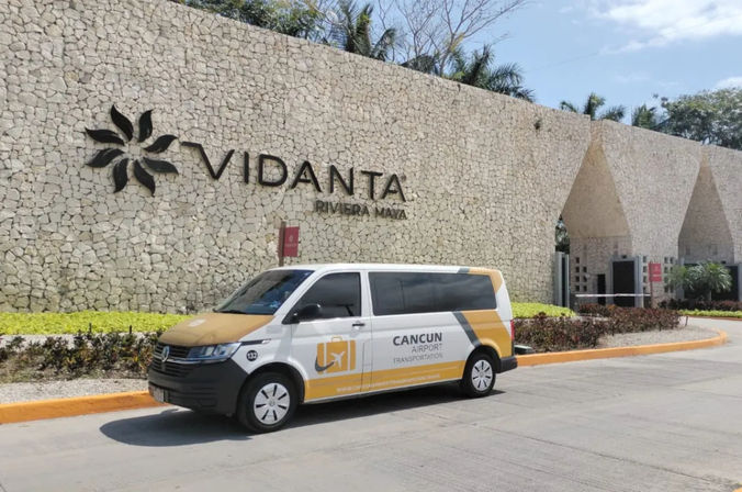 Yellow-and-white airport shuttle parked outside a Riviera Maya resort's textured stone entrance with a stylized flower logo, palm trees and sunny sky.