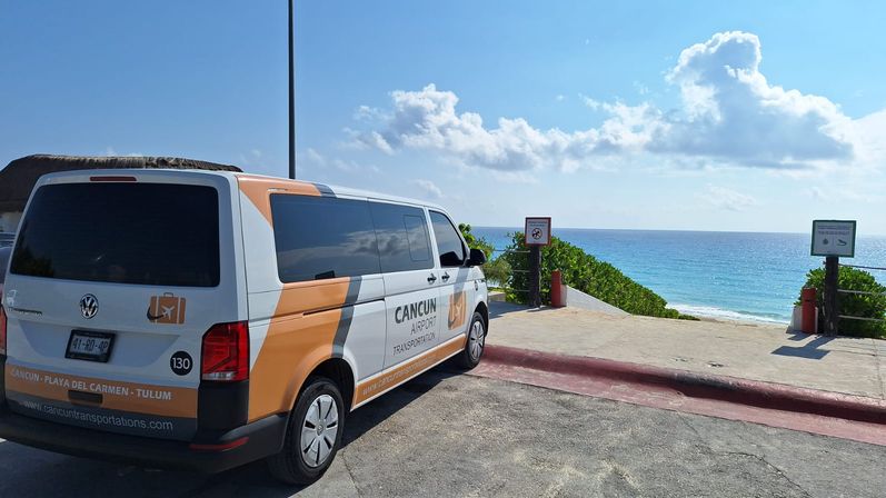 Orange-and-white airport shuttle van parked at a cliffside lookout overlooking turquoise Caribbean waters and a sandy shoreline in Cancún on a sunny day