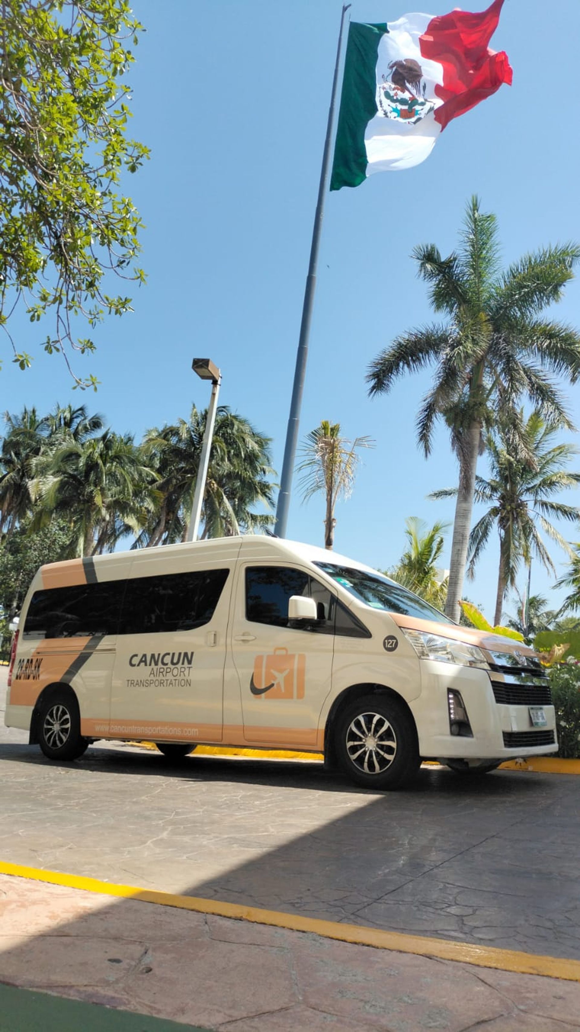White and orange airport shuttle van parked beneath a waving Mexican flag and tall palm trees on a sunny Cancun day.