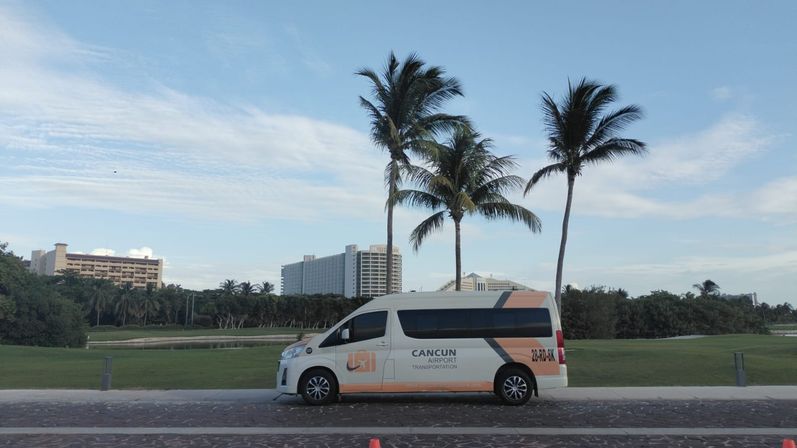 White airport shuttle van parked on a palm-lined coastal road in Cancun, Mexico, with resort hotels, green lawn and a bright blue sky.