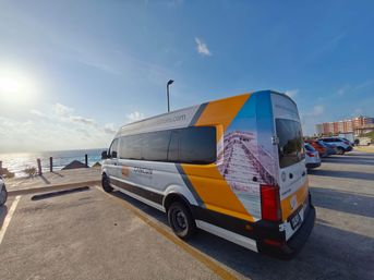 Sunlit shuttle van parked on a seaside lot in Cancun, Mexico with colorful Mayan pyramid graphics and ocean views at sunset