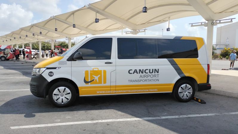White-and-yellow airport shuttle van parked under covered pickup bays at Cancun airport, luggage logo on the side and numbered curb signs visible