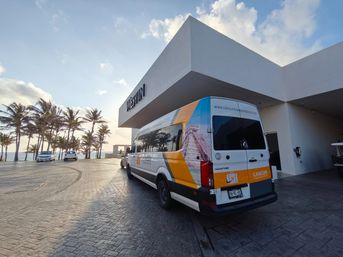 Bright shuttle van parked beneath a modern hotel canopy with palm-lined beachfront and ocean glowing at sunset in Cancún.