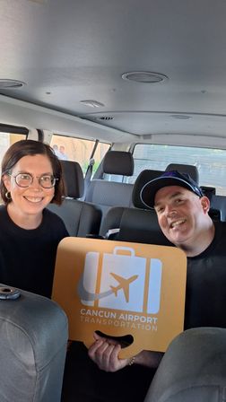 Two smiling travelers seated in a shuttle van holding an orange Cancun airport shuttle sign