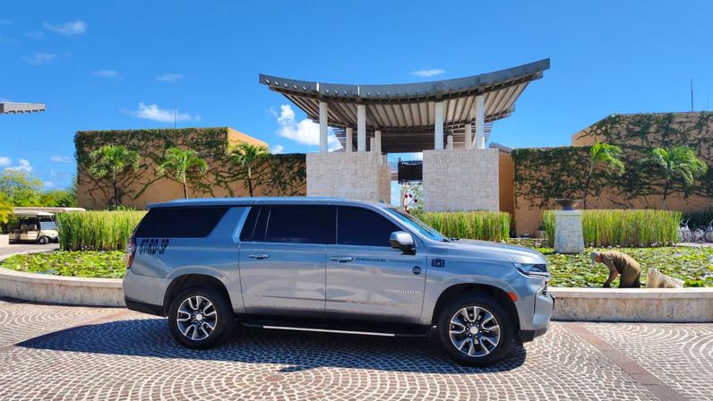 Silver SUV parked on a circular cobblestone driveway at a tropical resort entrance, with a lotus pond, palm trees, stone pavilion canopy, a gardener tending plants, and a bright blue sky