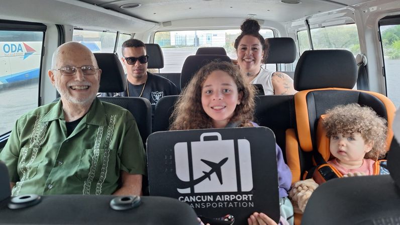 Multigenerational family smiling inside a Cancun airport shuttle van, a child holding a luggage sign with an airplane icon while a toddler rides in a car seat.