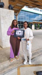 Two women smiling outside Cancun International Airport under a thatched canopy holding a black airport pickup sign for transportation.