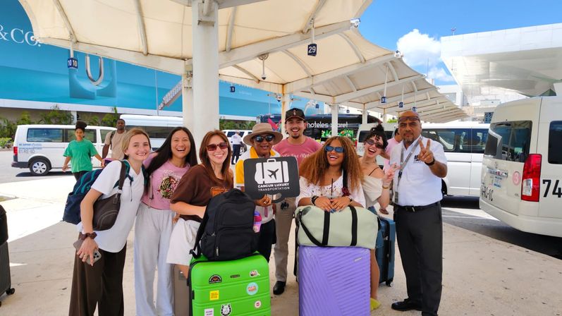 Smiling group of travelers with bright green and purple suitcases posing with shuttle staff under a shaded canopy at Cancun airport shuttle pickup