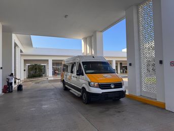 White-and-orange airport shuttle van parked under a sunny resort porte-cochère with travelers and luggage, decorative geometric columns and blue sky — Cancun resort drop-off.