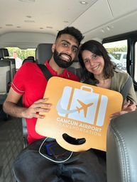 Smiling young couple inside a shuttle van holding an orange airport transfer sign with a suitcase and airplane icon, ready for a Cancún airport transfer.