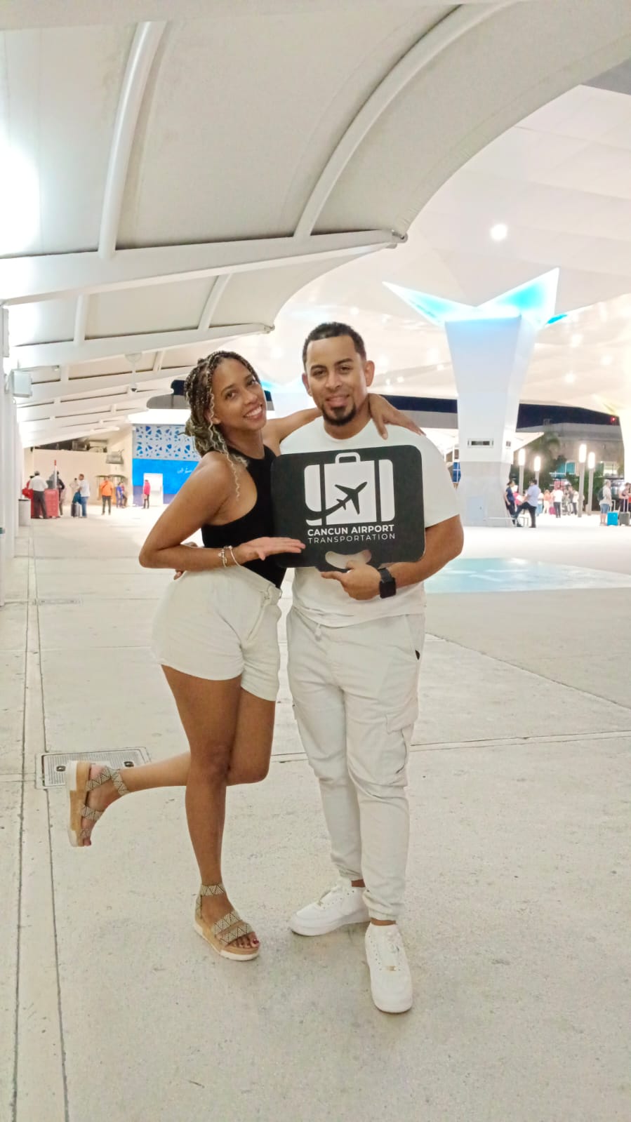 Smiling couple posing with an airport pickup sign in a modern Cancún airport terminal under illuminated canopy lighting