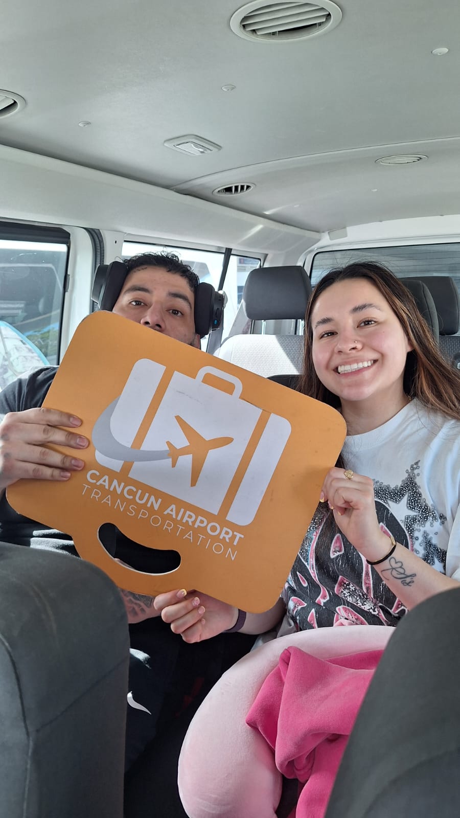 Smiling travelers in a shuttle hold an orange airport pickup sign with a suitcase and airplane icon labeled "Cancun".
