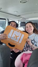 Smiling travelers in a shuttle hold an orange airport pickup sign with a suitcase and airplane icon labeled "Cancun".