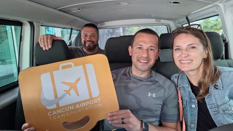 Three smiling travelers seated in an airport shuttle van holding a bright orange luggage-shaped sign with an airplane icon indicating airport transportation.