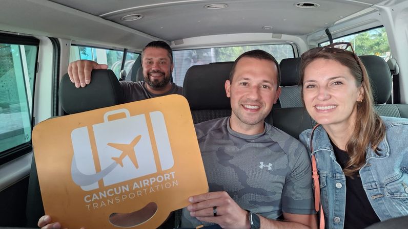 Three smiling travelers seated in an airport shuttle van holding a bright orange luggage-shaped sign with an airplane icon indicating airport transportation.