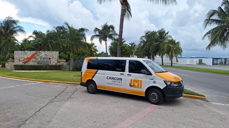 Yellow-and-white airport shuttle van parked by a tropical resort entrance with palm trees and ocean views in Cancún, Mexico under a cloudy sky.