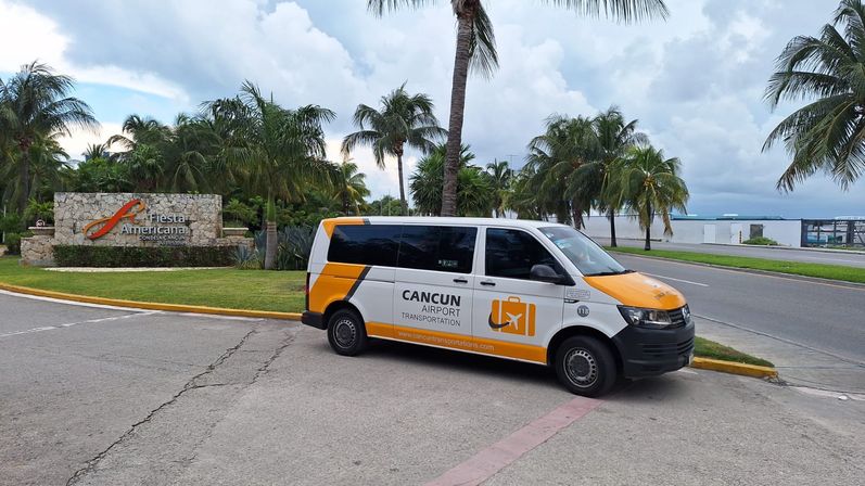 Yellow-and-white airport shuttle van parked by a tropical resort entrance with palm trees and ocean views in Cancún, Mexico under a cloudy sky.