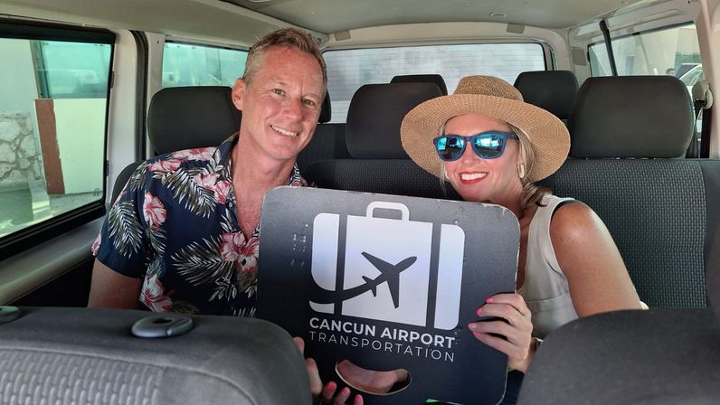 Smiling tourists in a shuttle van holding a Cancun airport shuttle sign — woman in a straw hat and blue sunglasses, man in a tropical print shirt, ready for vacation transfer.
