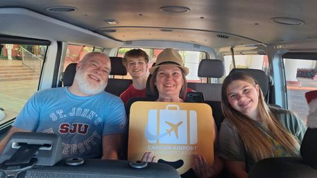 Four smiling passengers in a shuttle van holding an airport transfer sign with an airplane icon and 'Cancún' text, relaxed vacation vibe.