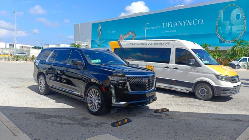 Gleaming black luxury SUV parked beside a white airport shuttle van in a sunny Cancún airport/resort parking lot with a large turquoise jewelry billboard in the background.