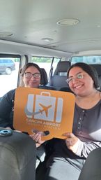 Two smiling passengers in a shuttle van holding an orange luggage-shaped sign with airplane icon for a Cancun airport shuttle transfer