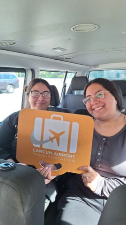 Two smiling passengers in a shuttle van holding an orange luggage-shaped sign with airplane icon for a Cancun airport shuttle transfer