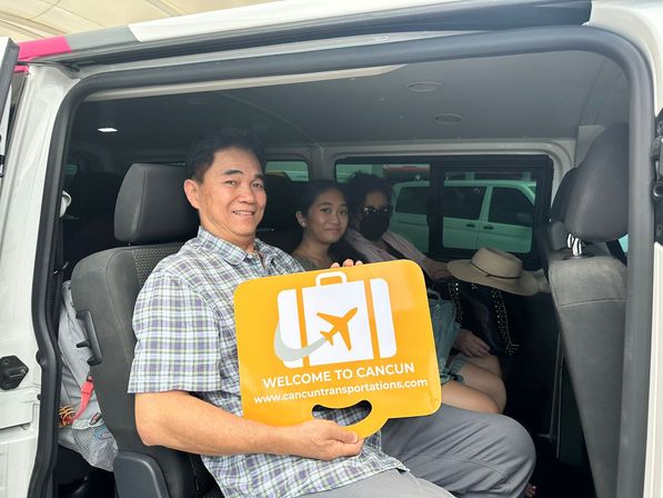 Smiling passengers seated inside a white airport shuttle van, man holding a bright orange "Welcome to Cancun" luggage sign — casual travelers arriving in Cancun.