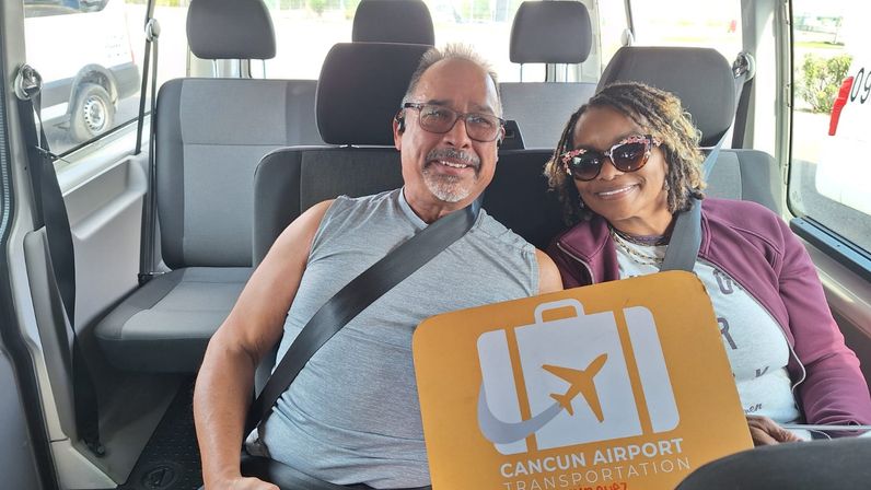 Happy couple buckled up in a shuttle van holding a bright Cancun Airport luggage sign with an airplane logo