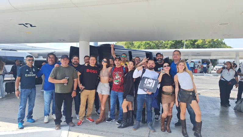 Cheerful group of travelers posing at an airport curbside by a black shuttle van under a covered canopy, casual summer outfits and boots, luggage visible — sunny shuttle pickup scene