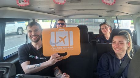 Cheerful group of four travelers inside a Cancun airport shuttle van, one holding an orange luggage sign with an airplane icon.