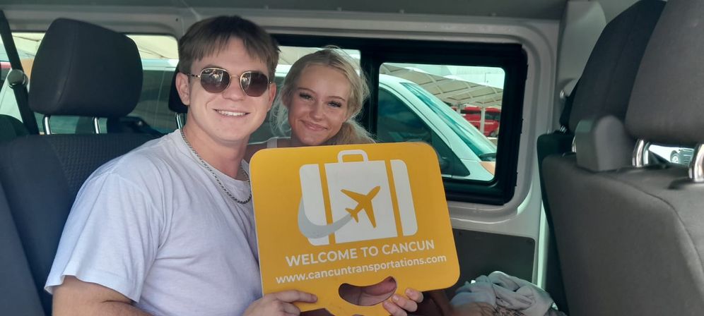 Smiling young couple seated in a shuttle van holding a bright yellow 'Welcome to Cancun' luggage sign, airport transfer vibe with parked cars visible through the window
