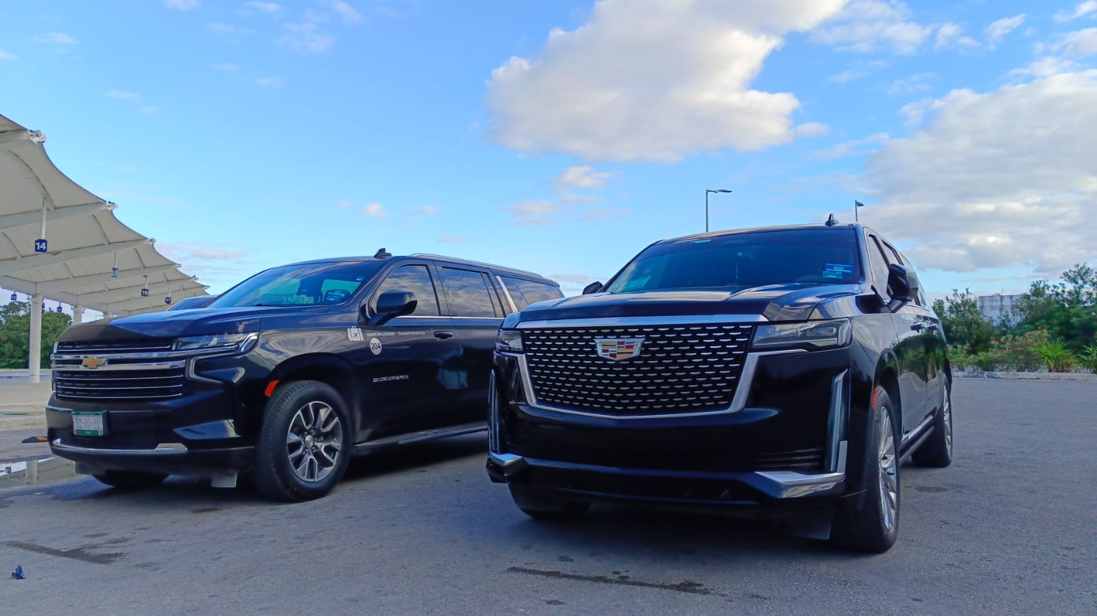 Two black full-size SUVs, a Cadillac Escalade and Chevrolet Suburban, parked side-by-side in an outdoor lot under a bright blue sky with scattered clouds